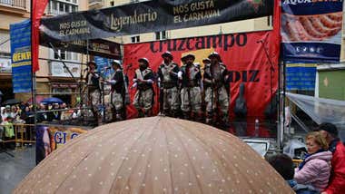 La comparsa 'Los sacrificaos' actúa en un tablao mientras el público se protege de la lluvia.