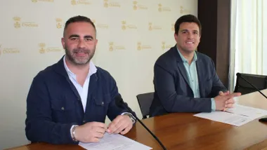 Fede Díaz y Salvador Guerrero sentados en una mesa de madera con el fondo en blanco con el escudo de Chiclana.
