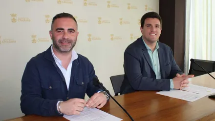 Fede Díaz y Salvador Guerrero sentados en una mesa de madera con el fondo en blanco con el escudo de Chiclana.