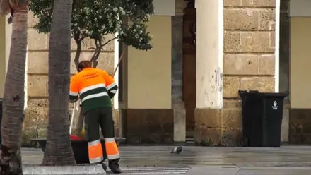 Mujer de la limpieza limpiando una calle, al fondo un edificio con un contenedor de basura.