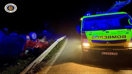 Vehículo rojo accidentado con las ruedas hacia arriba y camión de bomberos amarillo a su derecha, es de noche.