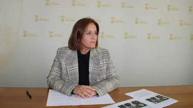 Ana González, teniente de alcalde de Chiclana, con papeles en una mesa de madera y fondo blanco con el escudo de Chiclana en amarillo-dorado.