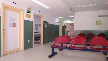 Sala de espera de un ambulatorio con asientos rojos, pared blanca y verde y puertas en tono blanco con marco beige.