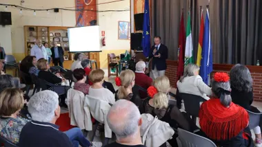 Salón de actos de San Antonio lleno de mayores que escuchan al alcalde de Chiclana, tras el el escenario y las banderas.