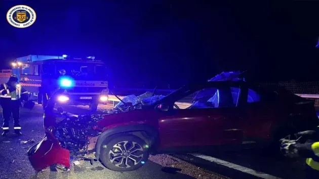 Coche rojo en la calzada con un fuerte impacto delantero, tras él los servicios de emergencia con las alarmas en azul.
