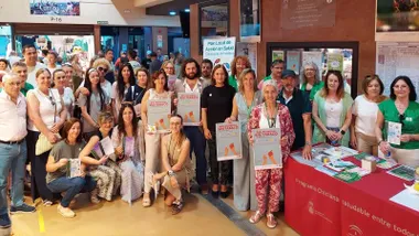 Muchas personas junto a la mesa, con tapete rojo, del Día Mundial Sin Trabajo, portan en sus manos el cartel de la misma.