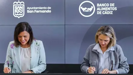 Patricia Cavada e Isabel Gomis en la firma del acuerdo entre el ayuntamiento de San Fernando y el Banco de Alimentos, tras ellas una pantalla en tonos grises.