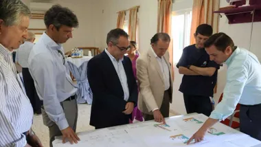 José María Román con chaqueta blanca, alcalde de Chiclana, mirando unos planos en una mesa y rodeado de otras personas.