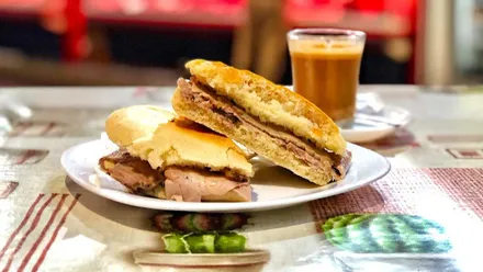 Un café junto a un mollete con lomo sobre un mantel con motivos de cocina.