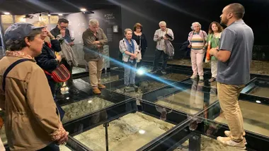 Un grupo de personas miran el suelo acristalado de Nueva Gadeira, bajo el cristal el yacimiento arqueológico.