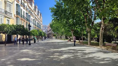 Un grupo de jóvenes camina por la Plaza de España de Cádiz mientras una mujer descansa en un banco