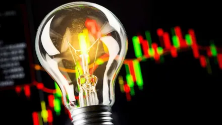 Bombilla con el filamento encendiéndose y tras ella un panel de la Bolsa en tonos rojos y verdes.