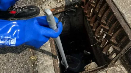 Operario con guantes azules buscando posibles nidos de mosquitos y larvas en una alcantarilla. Lo remueve con un extensor metálico.