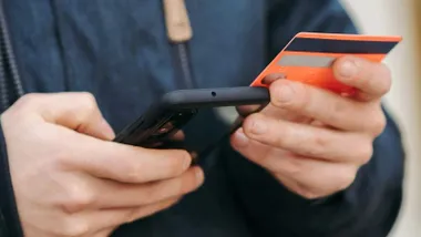 Hombre comprando a través del móvil, sostiene en su mano una tarjeta de crédito color naranja.