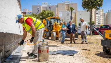 Trabajador pintando un muro en la zona ajardinada con responsables políticos tras de él, alejados.