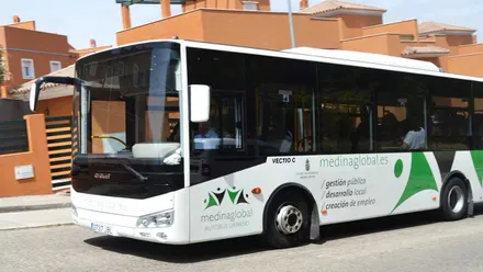 Autobús blanco con dibujos en verde de Medina Global, al fondo unos edificios de ladrillo.