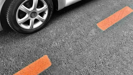 Asfalto pintado con rayas discontinuas en naranja, a la izquierda la rueda de un coche con chasis plateado.