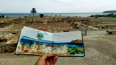 Mano sosteniendo un plano y al fondo las ruinas romanas de Bolonia, en Cádiz.