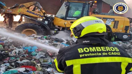Bombero con manguera apagando una montaña de residuos y retroexcavadora.
