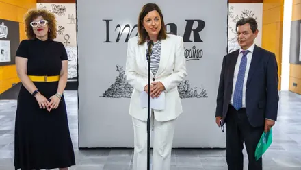 Patricia Cavada, con elegante traje blanco, en la presentación de "IMPAR", tras ella un cartel en blanco y negro de la exposición.