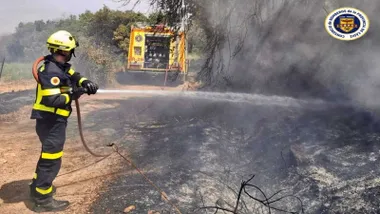 Bombero con una manguera apagando el incendio del terreno de trigal quemado y camión amarillo al fondo.