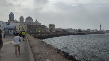 Cielos nublados en el Ca,po del Sur. varias personas pasean por el Malecón gaditano con la catedral y el Pirulí al fondo