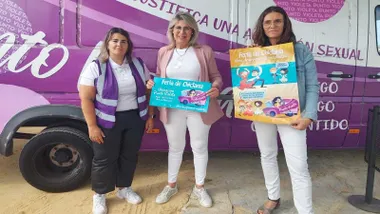 Responsables del Punto Violeta de la Feria de Chiclana portando carteles del mismo y tras ellas una furgoneta en colores blancos y violetas.