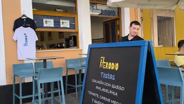Fachada del bar El Merodio en Cádiz. A la derecha, una antigua pizarra de tapas sin escribir y un camarero detrás. A la izquierda, dos camisetas con el logo del bar cuelgan de una ventana.