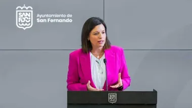 Patricia Cavada, alcaldesa de San Fernando, con chaqueta rosa y camisa blanca, tras ella panel gris y logotipo de San Fernando.