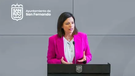 Patricia Cavada, alcaldesa de San Fernando, con chaqueta rosa y camisa blanca, tras ella panel gris y logotipo de San Fernando.
