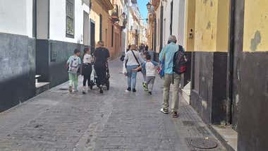 Varias personas, entre ellas niños y adultos, caminan por una calle al final del horario escolar. Algunos llevan mochilas.
