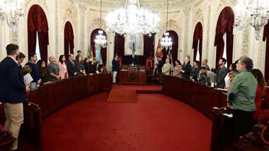 Vista general del salón de plenos del Ayuntamiento de Cádiz, con los miembros de la Corporación municipal de pie junto a sus escaños, distribuidos a ambos lados de la sala, bajo grandes lámparas de araña.