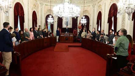 Vista general del salón de plenos del Ayuntamiento de Cádiz, con los miembros de la Corporación municipal de pie junto a sus escaños, distribuidos a ambos lados de la sala, bajo grandes lámparas de araña.
