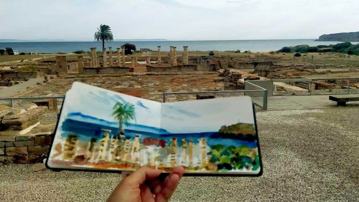 Mano sosteniendo un plano y al fondo las ruinas romanas de Bolonia, en Cádiz.