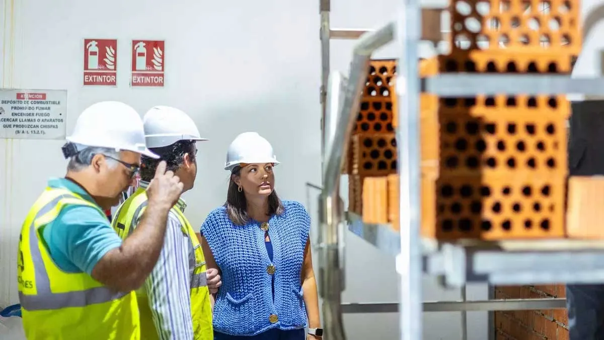 Patricia Cavada, con blusa azul, frente a un andamio con ladrillos y dos operarios que le muestran las obras, llevan chalequillos fluorescentes.