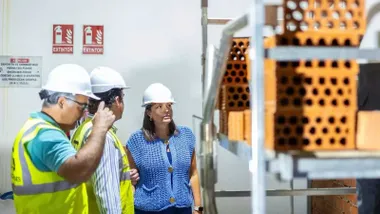 Patricia Cavada, con blusa azul, frente a un andamio con ladrillos y dos operarios que le muestran las obras, llevan chalequillos fluorescentes.