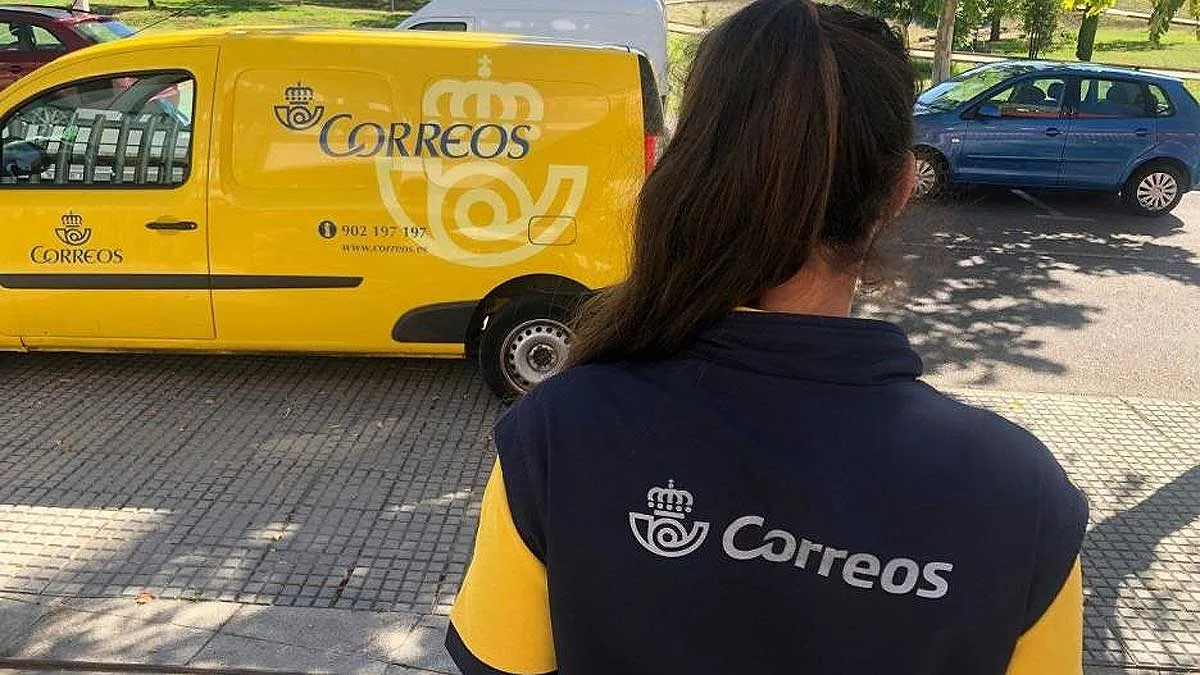 Empleada de Correos vestida de azul y amarillo y furgoneta al fondo.