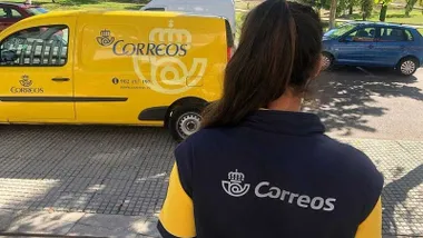Empleada de Correos vestida de azul y amarillo y furgoneta al fondo.