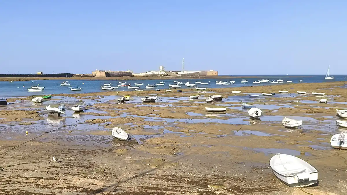 Espectacular bajamar en La Caletacon barquitas en primer término y el Castillo de San Sebastián al fondo en un día soleado con un cielo azul