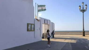 Una bandera de Andalucía ondea con fuerza por el Levante en una escuela infantil de Cádiz y una joven espera en la puerta
