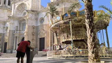 Hombre de espaldas bebiendo agua en una fuente tipo 'kichifuente' en la Plaza de la Catedral de Cádiz. Al fondo, se aprecian el tiovivo y la catedral bajo un cielo despejado.