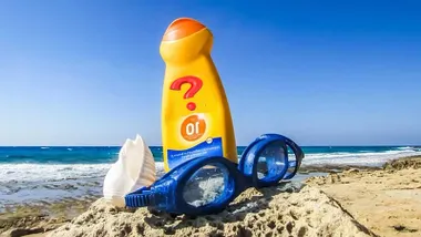 Bote de crema solar naranja en la playa junto a unas gafas de buceo en color azul.
