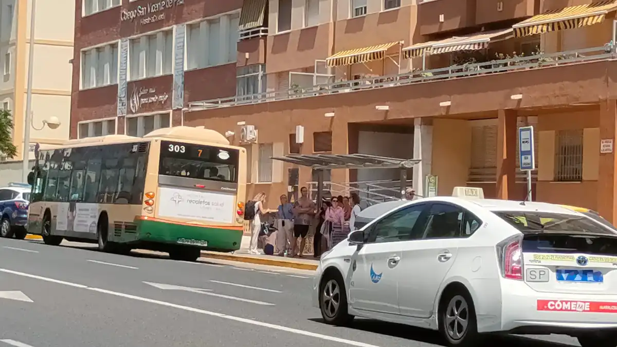 Autobús urbano de Cádiz lleno de pasajeros saliendo de una parada, mientras un taxi blanco espera detrás en la avenida