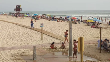 Vista de la playa de Cádiz un día de mar de fondo. Al fondo se distingue una torre de vigilancia con la bandera amarilla, que indica baño permitido con riesgo. En la orilla, varios bañistas disfrutan del agua mientras el cielo está despejado.