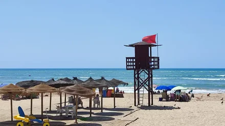 Una torre de vigilancia de socorrismo en la Playa de la Victoria de Cádiz la bandera roja que prohibe el baño izada y uan módulo de baño adaptado para personas con movilidad reducida en primer plano.