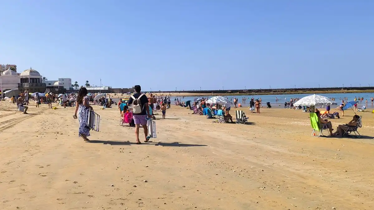 Fotografía tomada en la playa de La Caleta, en Cádiz, donde una pareja camina por la arena llevando sillas de playa. Al fondo, se aprecia el mar y parte de las murallas que rodean la playa. El cielo está despejado y la luz es intensa.