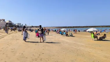 Fotografía tomada en la playa de La Caleta, en Cádiz, donde una pareja camina por la arena llevando sillas de playa. Al fondo, se aprecia el mar y parte de las murallas que rodean la playa. El cielo está despejado y la luz es intensa.