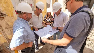 Bruno García y Virginia Martín observan los planos de rehabilitación en un edificio de la calle Soledad, con casco de obra y junto al cartel informativo del proyecto.