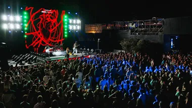 Escenario con un grupo cantando, pantallas gigantes en tonos rojos y luces blancas y verdes, hay mucho público.
