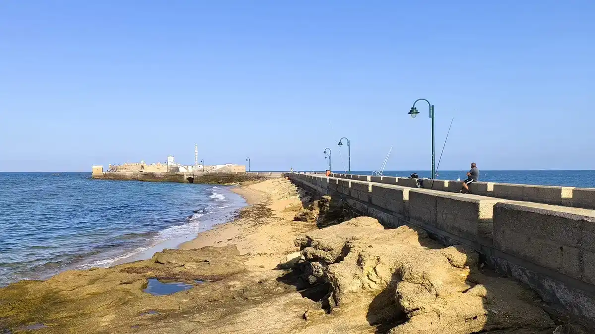 Vista del Castillo de San Sebastián en Cádiz al comienzo de la mañana, tomada desde las rocas junto al Paseo Quiñones. Se observan varias cañas de personas pescando. El cielo claro y la luz suave de la mañana.